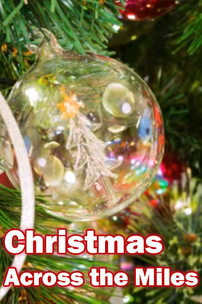 A closeup of a clear glass Christmas ball ornament on a Christmas tree. The lights from the tree reflect on its surface , and there is a little tree drawn on it. Christmas Across The Miles is written below it.